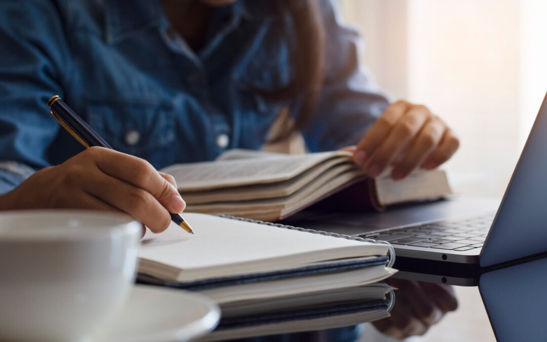 Concept photo with student holding book, in front of computer, with pen and notebook paper to signify keyboarding connecting to traditional literacy